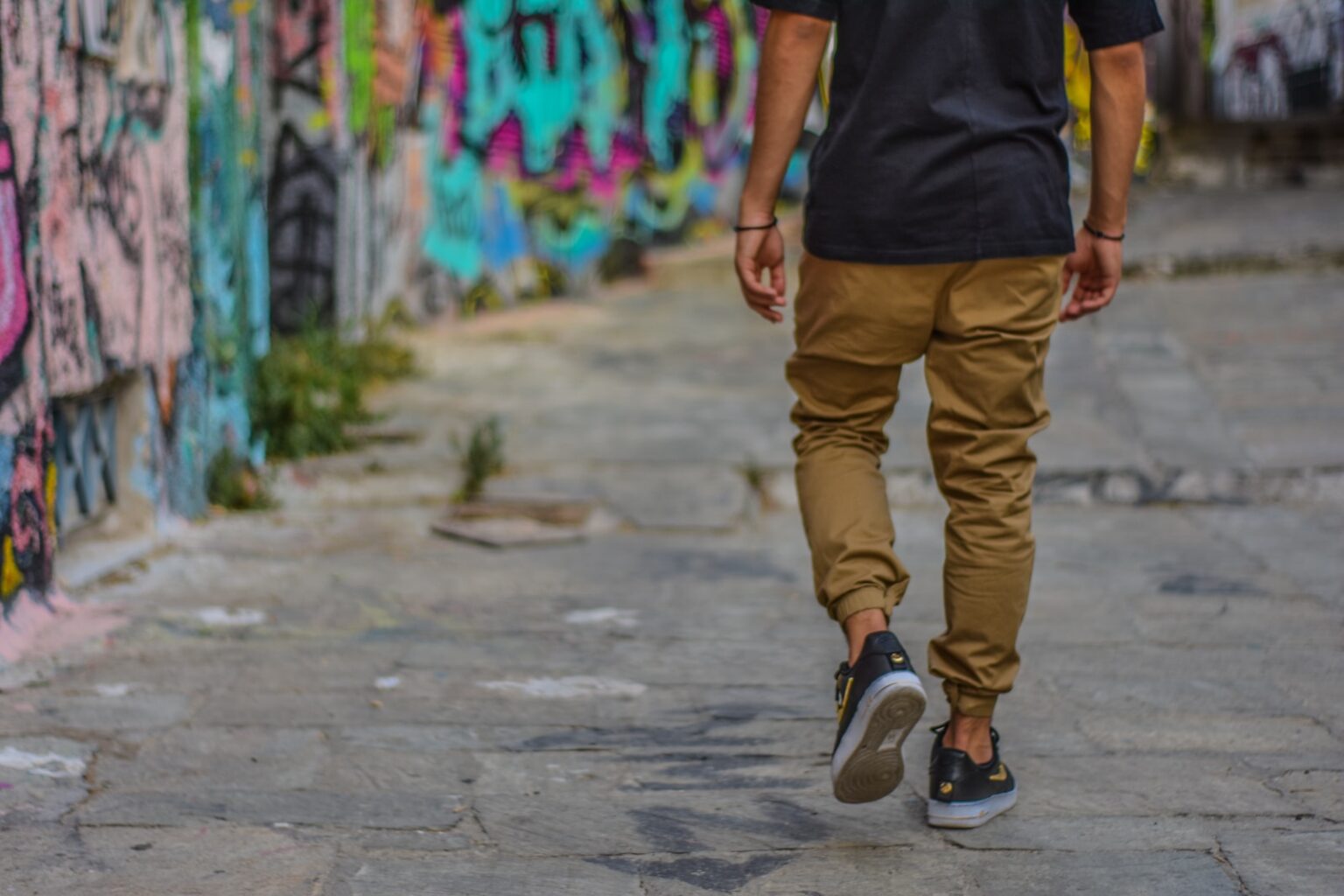 man wearing black shirt and brown pants walking beside wall with graffiti